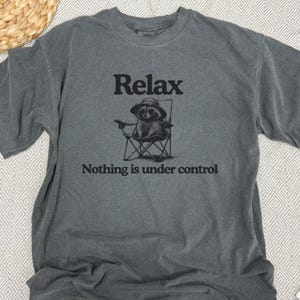 May include: Gray t-shirt with the words "Relax" and "Nothing is under control" printed in black. The shirt features a graphic of a raccoon wearing sunglasses and a hat, sitting in a folding chair.