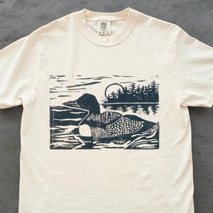 May include: Off-white t-shirt with a dark blue woodcut-style print of a loon on a lake, with trees and a sun. The shirt has a crew neck and short sleeves. The design is centered on the front of the tee.