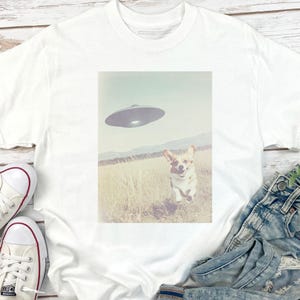 May include: White t-shirt with a graphic of a corgi running towards a UFO in a field. The graphic is in a vintage style.