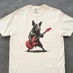 K&ouml;nnte beinhalten: Cremefarbenes T-Shirt mit einer schwarz-roten Grafik eines Bullterriers, der eine rote E-Gitarre spielt. Der Hund ist im Grunge-Stil dargestellt, wobei die Details der Gitarre sichtbar sind. Das Shirt hat einen klassischen Rundhalsausschnitt und kurze &Auml;rmel.