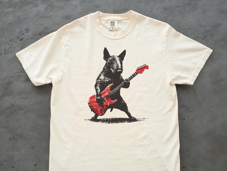 K&ouml;nnte beinhalten: Cremefarbenes T-Shirt mit einer schwarz-roten Grafik eines Bullterriers, der eine rote E-Gitarre spielt. Der Hund ist im Grunge-Stil dargestellt, wobei die Details der Gitarre sichtbar sind. Das Shirt hat einen klassischen Rundhalsausschnitt und kurze &Auml;rmel.