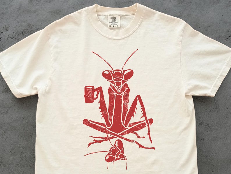 May include: Cream-colored t-shirt featuring a red graphic of a praying mantis holding a mug. The insect is depicted in a relaxed pose, with its legs crossed. The shirt has a crew neck and short sleeves.