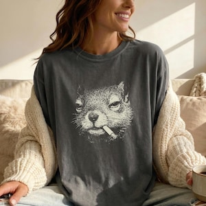 May include: Dark grey t-shirt with a black and white illustration of a squirrel smoking a cigarette. The squirrel's face is the main focus, with a slightly grumpy expression. The t-shirt has a relaxed fit.