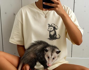 Pode incluir: Camiseta creme com uma ilustração em preto e branco de um gambá usando um chapéu de cowboy. Um gambá vivo está na frente da camiseta. A camiseta tem mangas curtas e gola redonda.