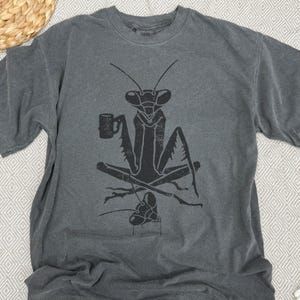 May include: Gray t-shirt featuring a black graphic of a praying mantis holding a mug. The mantis is seated with crossed legs, and a smaller mantis is below. The shirt has a crew neck and short sleeves.