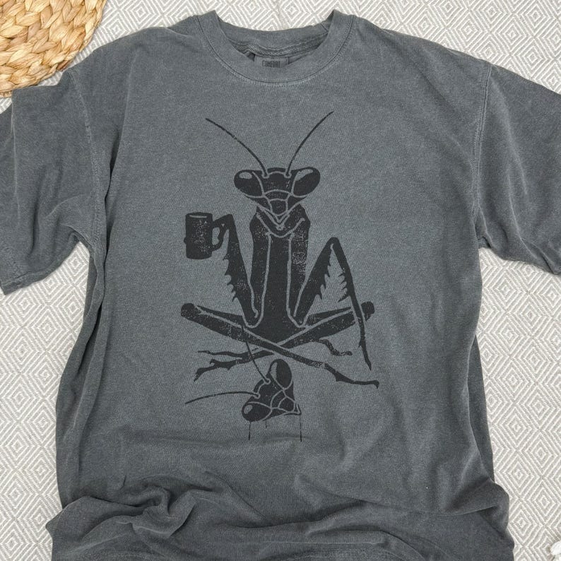 May include: Gray t-shirt featuring a black graphic of a praying mantis holding a mug. The mantis is seated with crossed legs, and a smaller mantis is below. The shirt has a crew neck and short sleeves.