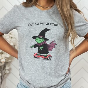 以下が含まれることがあります： 「Off to write code」の文字が入ったグレーのTシャツ。赤いホバーボードに乗った、魔女の帽子、スカーフ、マントを着たカエルの漫画が描かれています。カエルは緑色で、黒色のアクセントがあります。