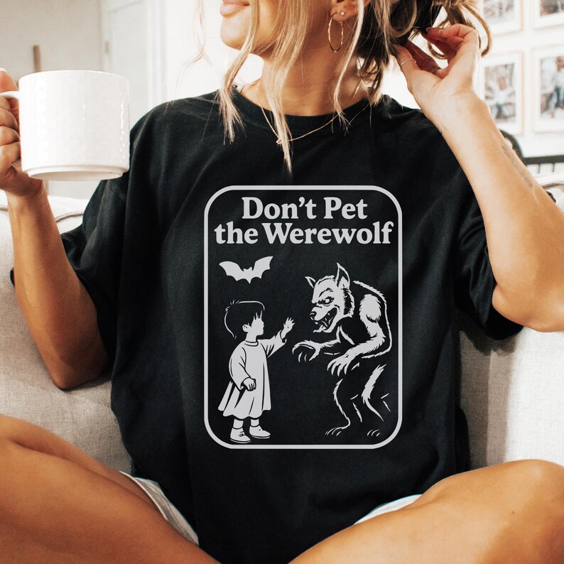 May include: Black t-shirt with a white graphic that reads "Don't Pet the Werewolf." The graphic features a child reaching toward a werewolf, with a bat above them. The shirt is a casual style.