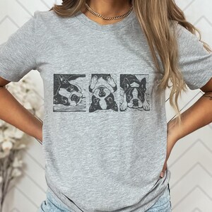 May include: A light gray t-shirt featuring a graphic of three Boston Terriers in black and white. The dogs are in different poses, each within a rectangular frame. The shirt has short sleeves and a crew neck.