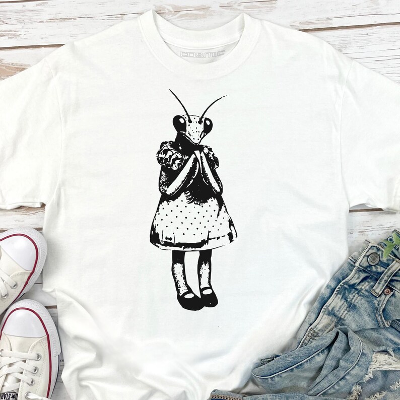 Puede incluir: Camiseta blanca con una ilustraci&oacute;n en blanco y negro de una mantis religiosa con un vestido de lunares. El insecto tiene las manos juntas. La camiseta se muestra con vaqueros azules y zapatillas blancas.