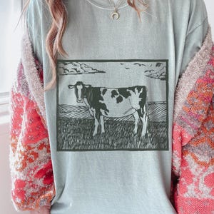 May include: A light blue t-shirt featuring a black and white illustration of a cow standing in a field under a cloudy sky. The cow has black spots and is facing forward. The shirt is a soft, comfortable material.