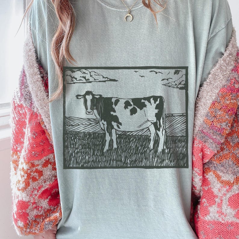May include: A light blue t-shirt featuring a black and white illustration of a cow standing in a field under a cloudy sky. The cow has black spots and is facing forward. The shirt is a soft, comfortable material.