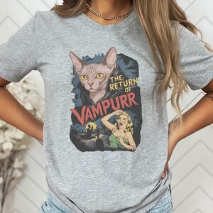 Peut inclure: T-shirt gris clair avec un motif graphique. Le motif repr&eacute;sente un chat avec des crocs de vampire et le texte "THE RETURN OF VAMPURR". Le motif comprend &eacute;galement une femme et une sc&egrave;ne de cimeti&egrave;re.
