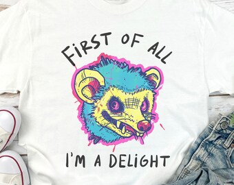 Puede incluir: Camiseta blanca con un gráfico de un opossum de dibujos animados en rosa, azul y amarillo. El texto "First of all I'm a delight" está impreso en la camiseta.