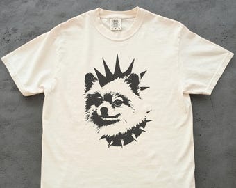 May include: Cream-colored t-shirt featuring a black graphic of a Pomeranian dog with a mohawk and spiked collar. The shirt has a crew neck and short sleeves. The design is a bold, punk-inspired illustration.