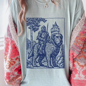 May include: Light blue t-shirt featuring a blue illustration of a knight riding a large dog, with a castle and tree in the background. The design has a vintage, woodcut style. The shirt is a comfortable, casual wear item.