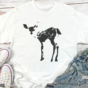 May include: White t-shirt featuring a black silhouette of a fawn. The fawn has a spotted pattern on its body. The shirt is paired with denim jeans and white sneakers. The shirt is made of a soft material.