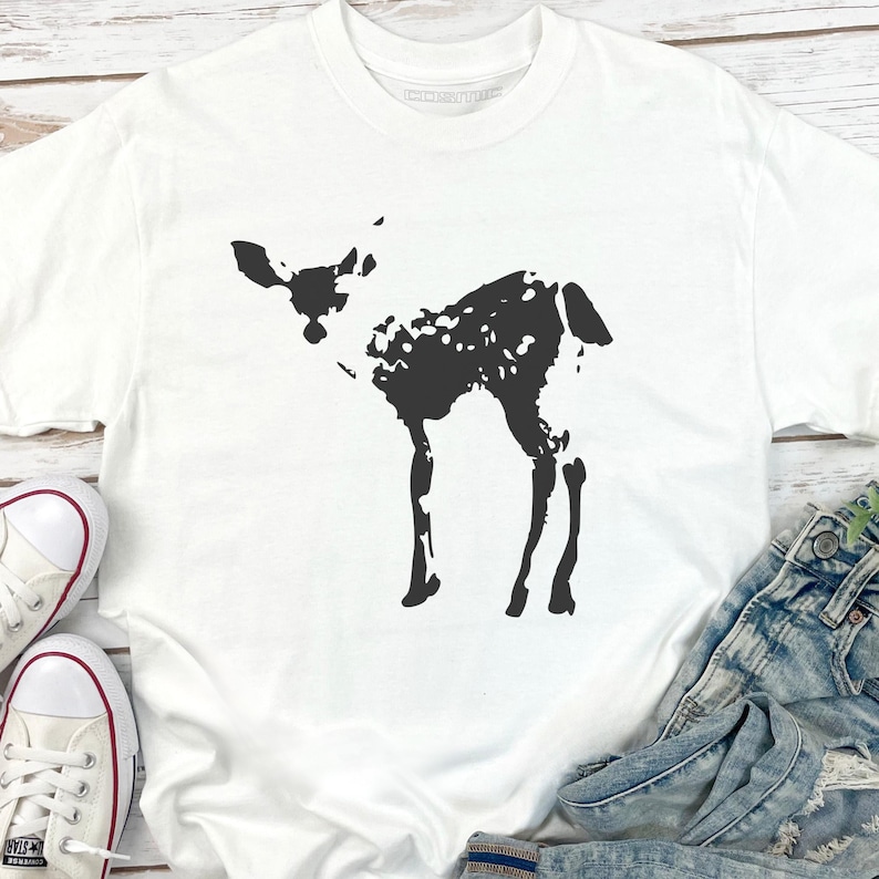 May include: White t-shirt featuring a black silhouette of a fawn. The fawn has a spotted pattern on its body. The shirt is paired with denim jeans and white sneakers. The shirt is made of a soft material.