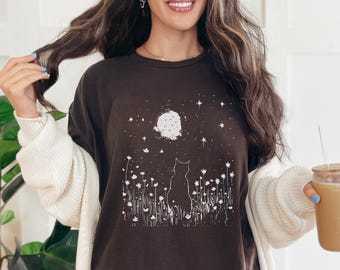 May include: A dark brown t-shirt featuring a white graphic of a cat silhouette gazing at a full moon and stars. The design includes stylized flowers. The shirt is part of a casual wear collection.