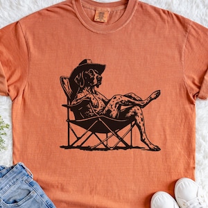 Puede incluir: Una camiseta de manga corta color naranja quemado con un estampado gráfico negro de un perro manchado con sombrero de vaquero, sentado en una silla de camping plegable con las piernas cruzadas. La prenda tiene cuello redondo y etiqueta.