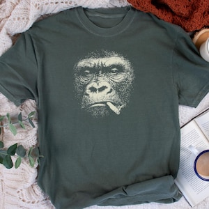 May include: Dark green t-shirt featuring a detailed, off-white illustration of a gorilla smoking a cigarette. The gorilla's face is the focal point, with a serious expression. The shirt is made of a soft material.