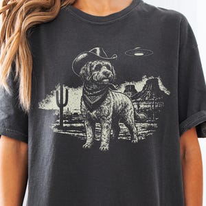 May include: A charcoal gray t-shirt featuring a graphic print of a dog wearing a cowboy hat and a bandana. The dog stands in a desert landscape with cacti, rock formations, and a flying saucer hovering in the sky above the scene.