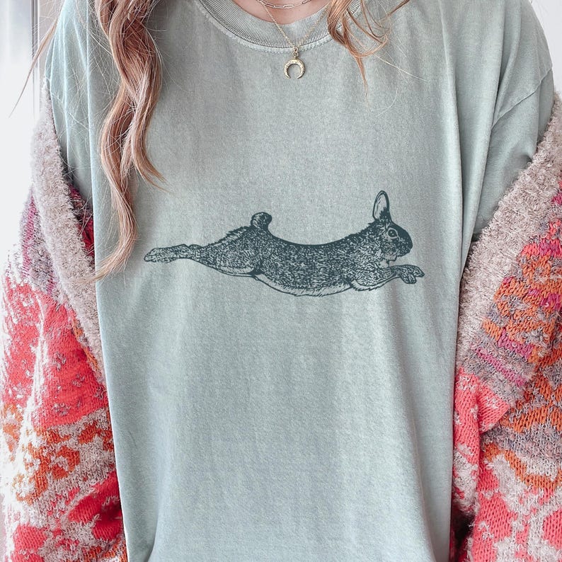 Puede incluir: Camiseta verde claro con una ilustraci&oacute;n gris oscura de un conejo corriendo. El conejo se muestra de perfil, con las orejas erguidas y las patas estiradas. La camiseta es de material suave y de corte holgado.