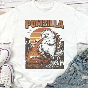 May include: White t-shirt with a graphic of a fluffy white dog labelled "Pomzilla" breathing fire at a car. The design includes a sunset, buildings, and a person with a dog. The shirt is paired with jeans and trainers.