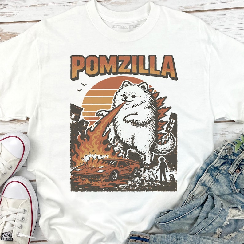 May include: White t-shirt with a graphic of a fluffy white dog labelled "Pomzilla" breathing fire at a car. The design includes a sunset, buildings, and a person with a dog. The shirt is paired with jeans and trainers.
