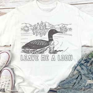 Puede incluir: Camiseta blanca con un gr&aacute;fico de un somorgujo nadando en un lago, con monta&ntilde;as y &aacute;rboles en el fondo. El texto "LEAVE ME A LOON" est&aacute; impreso debajo de la imagen. La camiseta es de material suave.