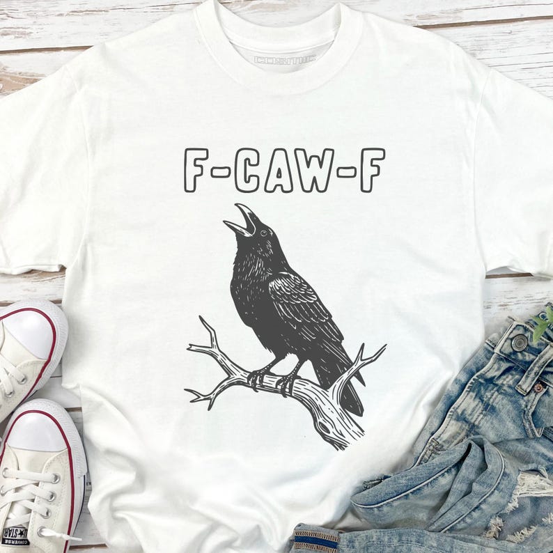May include: White t-shirt with a black graphic of a crow perched on a branch, with the text "F-CAW-F" above the bird. The crow is depicted with its beak open. The shirt is paired with denim jeans and white sneakers.