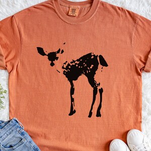 May include: Orange-colored t-shirt featuring a black silhouette of a deer. The shirt has short sleeves and a crew neck. The deer design is a simple, yet detailed illustration. The shirt is displayed on a white, textured surface.