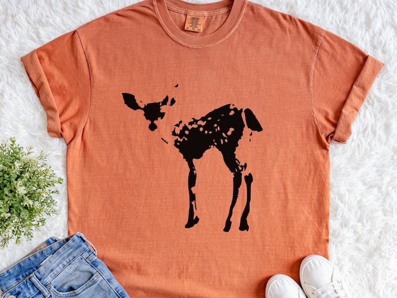 May include: Orange-colored t-shirt featuring a black silhouette of a deer. The shirt has short sleeves and a crew neck. The deer design is a simple, yet detailed illustration. The shirt is displayed on a white, textured surface.