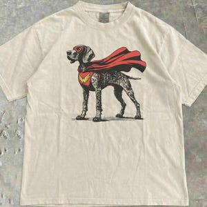 May include: Cream-colored t-shirt featuring a graphic of a dog in a superhero costume. The dog wears a red mask and cape, with a yellow emblem on its chest. The shirt has a classic crew neck and short sleeves.