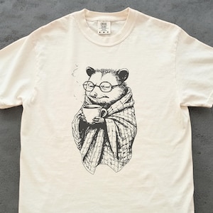 May include: Cream-colored t-shirt featuring a black and white illustration of an opossum wearing glasses and a blanket, holding a cup of coffee. The opossum is depicted with steam rising from the cup. A casual, comfortable tee.