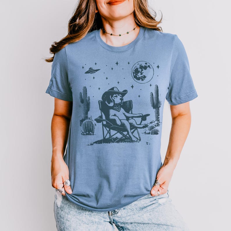 Pu&ograve; includere: Una maglietta blu chiaro con una grafica di un cane che indossa un cappello da cowboy e siede su una sedia da campeggio sotto una luna crescente e stelle. Il cane &egrave; circondato da cactus e un UFO.