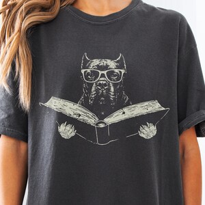 May include: A charcoal grey short-sleeve t-shirt featuring a graphic print of a dog wearing glasses and reading an open book. The illustration is rendered in a light cream colour, contrasting against the dark fabric of the crew neck shirt.