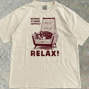 May include: Cream-colored t-shirt with a maroon graphic of a dog sleeping in a striped armchair. The text "NOTHING IS UNDER CONTROL!" and "RELAX!" are above and below the image.