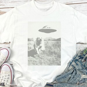 May include: White t-shirt with a graphic of a dog looking up at a flying saucer. The graphic is in black and white and the background is a field of grass.