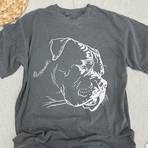 Könnte beinhalten: Ein dunkelgraues T-Shirt mit einer weißen Umrandung eines Hundekopfes. Der Hund hat einen ernsten Ausdruck und schaut nach rechts.