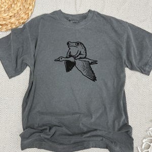 K&ouml;nnte beinhalten: Ein anthrazitfarbenes Kurzarm-T-Shirt mit einem schwarzen Grafikdruck, der einen Frosch zeigt, der auf dem R&uuml;cken einer fliegenden Gans sitzt. Das T-Shirt hat einen Rundhalsausschnitt und eine lockere Passform mit einer detaillierten Illustration.