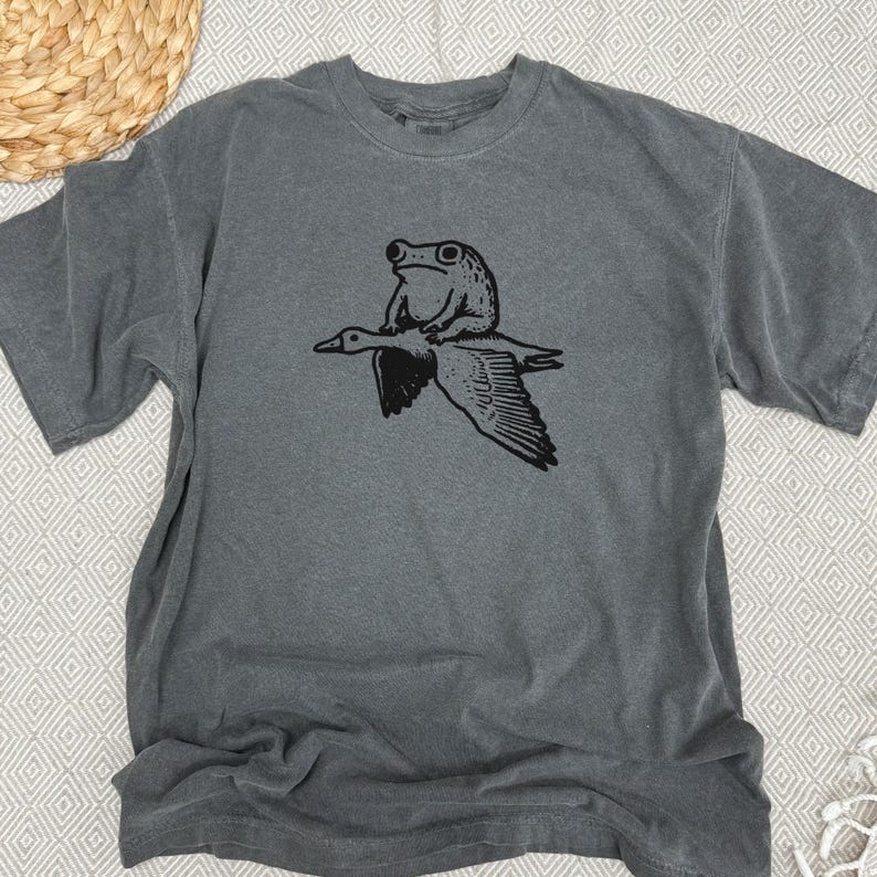 K&ouml;nnte beinhalten: Ein anthrazitfarbenes Kurzarm-T-Shirt mit einem schwarzen Grafikdruck, der einen Frosch zeigt, der auf dem R&uuml;cken einer fliegenden Gans sitzt. Das T-Shirt hat einen Rundhalsausschnitt und eine lockere Passform mit einer detaillierten Illustration.
