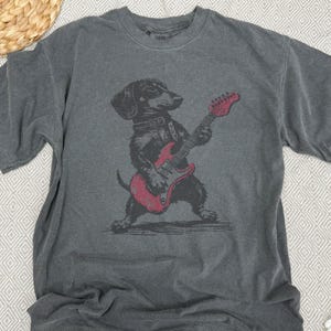K&ouml;nnte beinhalten: Graues T-Shirt mit einer schwarz-roten Illustration eines Dackels, der eine rote E-Gitarre spielt. Der Hund tr&auml;gt ein Halsband. Das Shirt hat kurze &Auml;rmel und einen Rundhalsausschnitt.