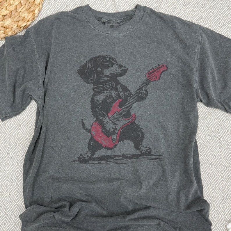 K&ouml;nnte beinhalten: Graues T-Shirt mit einer schwarz-roten Illustration eines Dackels, der eine rote E-Gitarre spielt. Der Hund tr&auml;gt ein Halsband. Das Shirt hat kurze &Auml;rmel und einen Rundhalsausschnitt.