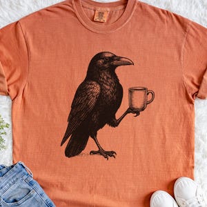 Op de afbeelding: Oranje T-shirt met een zwarte illustratie van een kraai die een koffiemok vasthoudt. De kraai is getekend in een vintage stijl. Het shirt is gemaakt van zacht materiaal en heeft een ronde hals. Het ontwerp is gedrukt op een comfortabel, casual T-shirt.