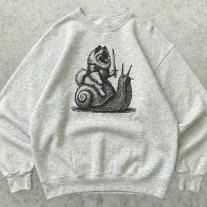 Puede incluir: Una sudadera de cuello redondo en color gris jaspeado claro con un estampado gráfico negro estilo tinta que muestra a un perro peludo con armadura y una espada montado sobre el lomo de un caracol grande. La prenda tiene mangas largas.