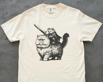 May include: Off-white t-shirt with a black and white illustration of a cat and frog. The cat has a stern expression, and the frog, wearing a cape, is holding a sword. The t-shirt has a crew neck and short sleeves. The fabric appears to be cotton.