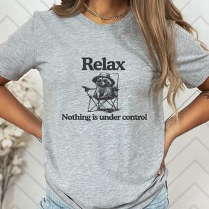 May include: A heather gray t-shirt featuring the word "Relax" above an illustration of a raccoon in a folding chair. The text "Nothing is under control" is below the illustration. The shirt is a casual wear item.