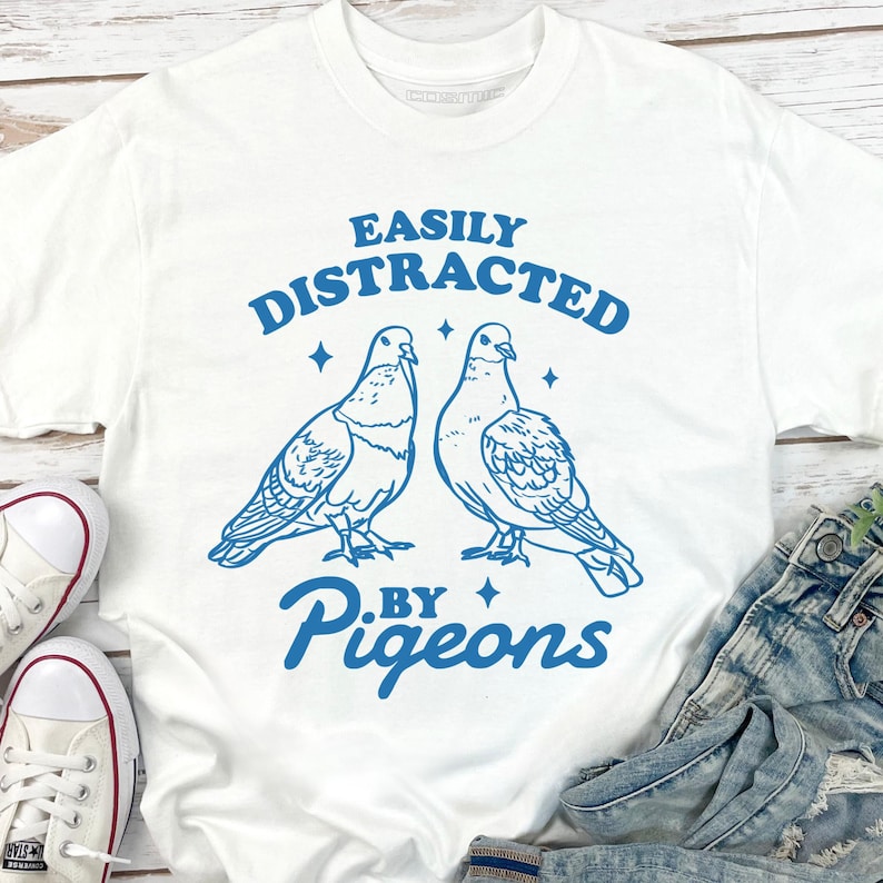 May include: White t-shirt with a blue graphic design featuring two pigeons and the text "EASILY DISTRACTED BY PIGEONS". The design includes stars and a playful font. The shirt is paired with denim jeans and white trainers.