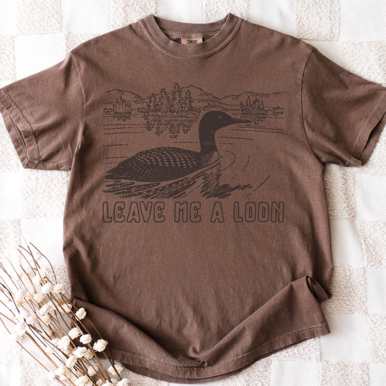 Puede incluir: Camiseta marr&oacute;n con un dibujo de un somorgujo nadando en un lago, con monta&ntilde;as al fondo. El texto "LEAVE ME A LOON" est&aacute; impreso debajo. La camiseta es de un material suave.